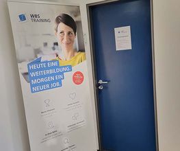 WBS TRAINING Werbeaufsteller mit der Aufschrift „Heute eine Weiterbildung. Morgen ein neuer Job.“ vor einer blauen Tür..