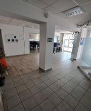 Eingangsbereich mit Säule. Links ein Regal mit Pflanzen und ein rotes Sofa, rechts eine Glaswand. Im Hintergrund sind mehrere Arbeitsplätze erkennbar.