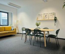 Pausenraum mit langem Tisch und 6 Stühlen. Links ein gelbes Sofa und eine Stehlampe. An der Wand ein Regal mit 3 Bildern und einer Zimmerpflanze.