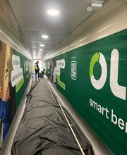 mediaService Bremen | Full-Stack Werbeagentur - Zwei Werbetechniker bei der Montage einer großflächigen grünen Wandverkleidung mit Werbeaufdrucken und QR-Code in einem langen Korridor.