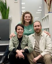 3 Bildungsberater:innen sitzen auf einer Treppe und schauen freundlich in die Kamera. Im Hintergrund sieht man eine große Grünpflanze, ein Büro und ein weißes Regal.