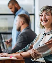 Eine Frau sitzt vor einem Laptop und blickt in die Kamera. Im Hintergund sind unscharf zwei Männer zu sehen.