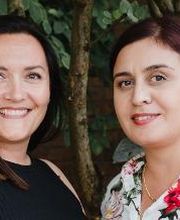 Bild von 2 Mitarbeiterinnen die vor einem grünen Strauch stehen und freundlich in die Kamera lächeln.