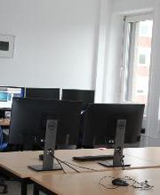 Schulungsraum mit Schreibtischen auf denen sich jeweils zwei Bildschirme, eine Tastatur, Maus und Headset befinden. In dem Raum befindet sich eine Person, die lernt. Fensterfront.