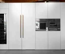 Elegante, moderne Hochschrankküche mit großflächigen weißen Fronten, integrierten schwarzen Einbaugeräten und einer schlanken Glasfront. Die grifflosen Schrankelemente mit langen Holzgriffen verbinden minimalistisches Küchendesign mit warmen natürlichen Akzenten. Die sichtbare Holzbalkendecke ergänzt die hochwertige, zeitlose Küchengestaltung.