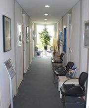 Flur mit Stühlen und Tischen sowie einem großen Fenster vor dem eine grüne Zimmerpflanze steht.