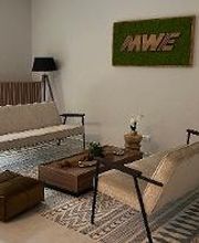 Foyer mit 2 modernen und stilvollen Sofas sowie einem braunen Beistelltisch und einem braunen Sitzkissen. Im Hintergrund eine Stehlampe sowie eine grüne Zimmerpflanze vor einem großen Fenster.