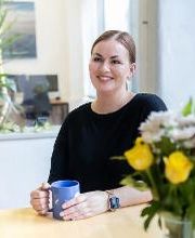 Frau mit braunem Haar sitzt am Tisch und hält eine blaue Tasse. Vor ihr steht ein Strauß Blumen. Im Hintergrund befinden sich Arbeitsplätze und Pflanzen.