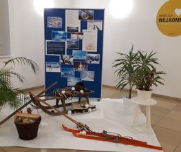 Winter-Dekoration im Foyer. Ein Schlitten, Skier und ein Hockeyschläger auf einem weißen Teppich. Dahinter ein Aufsteller mit verschiedenen Fotos und einigen Zimmerpflanzen. An der Wand hängt ein gelber Aufkleber mit der Aufschrift Herzlich Willkommen.