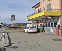 AGROLA Tankstelle in Kradolf-Schönenberg; vor Tankstelle weisses Auto; über Tankstellen-Dach Balkon; links Totem mit AGROLA Schild und digitaler Preisanzeige; dahinter Landwirtschafts-Feld