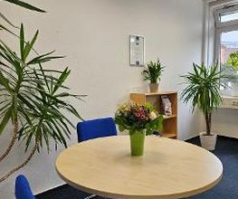 Ein heller Raum mit einem runden Holztisch, darauf ein Blumenstrauß in einer grünen Vase. Um den Tisch stehen blaue Stühle. Im Hintergrund stehen Grünpflanzen, ein kleines Regal und ein Fenster, das Tageslicht hereinlässt. Die Wände sind weiß.