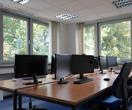 Mehrere Arbeitsplätze mit jeweils 2 Bildschirmen, einem Bürostuhl, Tastatur, Maus und Headset. Raum mit vielen Fenstern und Blick ins Grüne. 