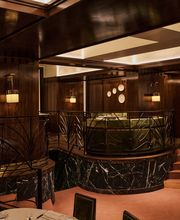View of a multi-level restaurant interior featuring dark marble bases, intricate iron railings, green velvet banquettes, warm wood walls, and pink carpeting.