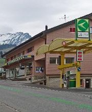 AGROLA Tankstelle in Amden. Gelbe Glasüberdachung mit einer Zanksäule und Zahlterminal. Digitaler Preisanzeige. Dahinter rotes Gebäude mit Volg und Bergkette im Hintergrund der Aufnahme.