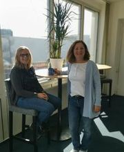 Bild von 2 Mitarbeiterinnen, die am Hochtisch vor dem Fenster stehen und freundlich in die Kamera lachen. Auf dem Tisch steht eine grüne Zimmerpflanze.