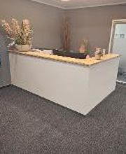 Empfangsbereicht mit modernem Thresen, 2 Sesseln, einem großen Fernseher auf dem ein standbild mit Informationen zu sehen ist. Mehrere Topfpflanzen. Blick in den Flur.