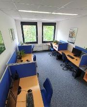 Schulungsraum mit mehreren Arbeitsbereichen, die durch blaue Trennwände voneinander abgegrenzt sind und mit 2 PC-Bildschirmen, Tastatur, Maus und Headset ausgestattet sind.