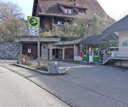 AGROLA Tankstelle in Maschwanden; zwei Tanksäulen zwischen Gebäuden gelegen. Links Pfeiler mit AGROLA Logo und Preisschildern