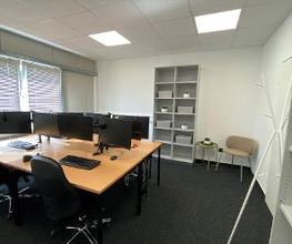 Schreibtische im Trainingsraum mit jeweils zwei PC-Bildschirmen, Tastatur, Maus und Headset. Im Hintergrund steht ein Bücherregal. Daneben ist eine Sitzgelegenheit mit einem Beistelltisch