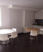 Ein Pausenraum mit zwei runden Tischen und weißen Stühlen. Rechts ist eine Küchenzeile mit weißen Schränken, Kaffeemaschinen und einer Mikrowelle. Ein großes Fenster lässt Tageslicht herein, und der Boden ist dunkelgrau.