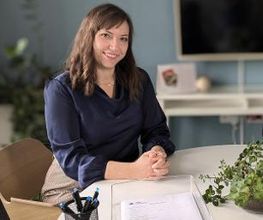 Frau sitzt am Tisch. Vor ihr eine Ablage mit Zetteln und mehrere Stifte. Im Hintergrund ein TV