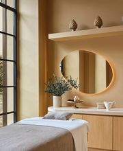 Calming spa treatment room with a massage bed, circular mirror, and built-in shelving decorated with natural elements at Aelia Spa.