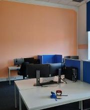 Arbeitsbereich mit 2 Computer-Bildschirmen, einer Tastatur, Maus und blauen Trennwänden. Auf dem Tisch liegen 2 Locher und links befindet sich ein Garderobenständer. 