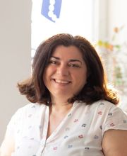 Auf dem Foto ist Denise Kir zu sehen, die Teamassistentin bei WBS TRAINING. Sie lächelt freundlich in die Kamera, trägt eine helle Bluse mit buntem Muster und sitzt in einem hellen Raum. Ihr Ausdruck wirkt warm und einladend, was ihre Rolle als unterstütz