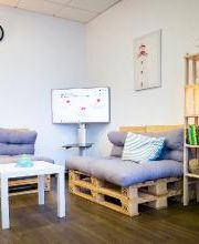 Gemütliche Sitzecke aus 2 modernen Palettensofas mit grauen Kissen, einer Stehlampe, einem Regal und einem Fernseher. Im Hintergrund ein Bild von einem Leuchtturm.