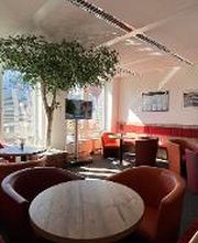 Pausenbereich mit mehren Tischen und roten Sesseln. Große Fensterfront vor der eine große Zimmerpflanze steht. An der Wand hängen Bilder und Informationen.