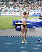 Eine Leichtathletin steht im Stadion auf der Laufbahn und hebt beide Arme in einer Geste der Konzentration oder des Abschlusses. Die Szene wirkt ruhig und zugleich wettkampforientiert.