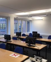 Ein Raum mit 3 Tischreihen an denen. blaue Stühle stehen. Auf den Tischen befinden sich Bildschirme sowie Tastatur und Maus. An der Seite des Raumes befindet sich eine Fensterfront mit Blick ins Grüne.