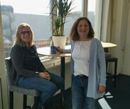 Bild von 2 Mitarbeiterinnen, die am Hochtisch vor dem Fenster stehen und freundlich in die Kamera lachen. Auf dem Tisch steht eine grüne Zimmerpflanze.