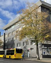 Außenansicht vom Gebäude an einer Straße. Graue Steinfassade. Gelbe Busse an der Straße. Schildertafel im Eingangsbereich.