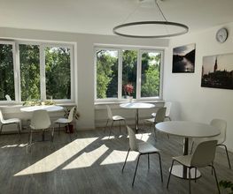 Ein heller Aufenthaltsraum mit großen Fenstern mit Blick ins Grüne. In dem Raum befinden sich runde, weiße Tische und passende Stühle. An der Wand hängen Leinwandbilder von Hamburg.
