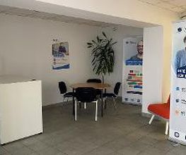 Eingangsbereich mit einer Theke, einem Tisch mit 4 schwarzen Stühlen und 2 Roll up Banner. An der Wand ein Plakat und rechts ein roter Stuhl.
