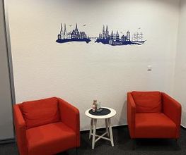 Ein kleiner Sitzbereich mit zwei roten Sesseln und einem weißen Beistelltisch dazwischen. Auf dem Tisch steht eine kleine Dekoration. An der Wand ist eine blaue Silhouette von Lübeck mit dem Schriftzug „Lübeck“ angebracht.