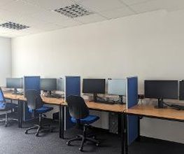 Schulungsraum mit mehreren Arbeitsbereichen, die durch blaue Trennwände voneinander abgegrenzt sind und mit PC-Bildschirmen, Tastatur, Maus und Headset ausgestattet sind. Blaue Bürostühle.
