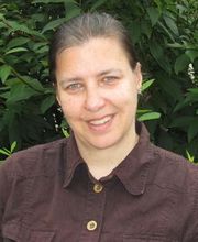 Frau mit braunem Haar steht draußen vor grünen Pflanzen und schaut freundlich in die Kamera.