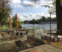 Bild von der Terrasse mit Sitzgelegenheiten und Sonnenschirmen. Daneben ein Gewässer.