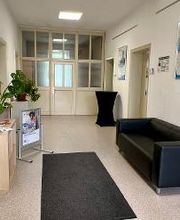 ein Eingangsbereich mit einer schwarzen Couch auf der rechten Seite. Links steht ein Regal mit Flyern und einer Zimmerpflanze. Am Ende des Raums steht ein Stehtisch neben einer Glastür. Auf dem Boden liegt ein Teppich. 