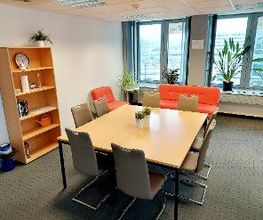 Pausenraum mit großem Tisch mit 8 grauen Stühlen. Links steht ein Holzregal mit Büchern und Deko, im Hintergrund ein rot/oranges Sofa und ein Sessel. Auf dem Fensterbrett stehen mehrere Zimmerpflanzen.