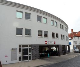 Außenansicht des Gebäudes, Halbrunde Form und helle Fassade. Im Erdgeschoss befindet sich ein Parkhaus.