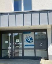 Eingangsbereich an einem hellen, sonnigen Tag. Graue Überdachung und Zitronengelbe Wand. Parkmöglichkeiten.