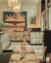 Elegant café interior with marble pastry counters, illuminated display cases of baked goods, sculptural pendant chandeliers, and neatly set tables with white linens and single-stem floral arrangements.