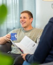 Mann sitzt in einem Sessel und hält eine blaue Tasse in der Hand. Ihm gegenüber sitzt eine Frau mit einem Flyer in der Hand.