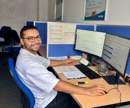 Teilnehmer sitzt am Arbeitsplatz mit 2 Bildschirmen, Tastatur, Maus und Zetteln. Im Hintergrund befinden sich weitere Arbeitsplätze.