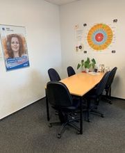 Büro bestehend aus einem langen Tisch mit mehreren schwarzen Bürostühlen. An der Wand hängen Plakate und auf dem Tisch steht eine kleine Topfpflanze.