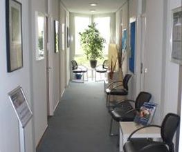 Flur mit Stühlen und Tischen sowie einem großen Fenster vor dem eine grüne Zimmerpflanze steht.