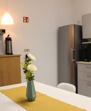 Küche mit mehreren Schränken, Wasserkocher, Kaffeemaschine, Wassersprudler und Ofen. Im Vordergrund ein Tisch mit mehreren Stühlen und darauf einer Blume in einer blauen Vase..